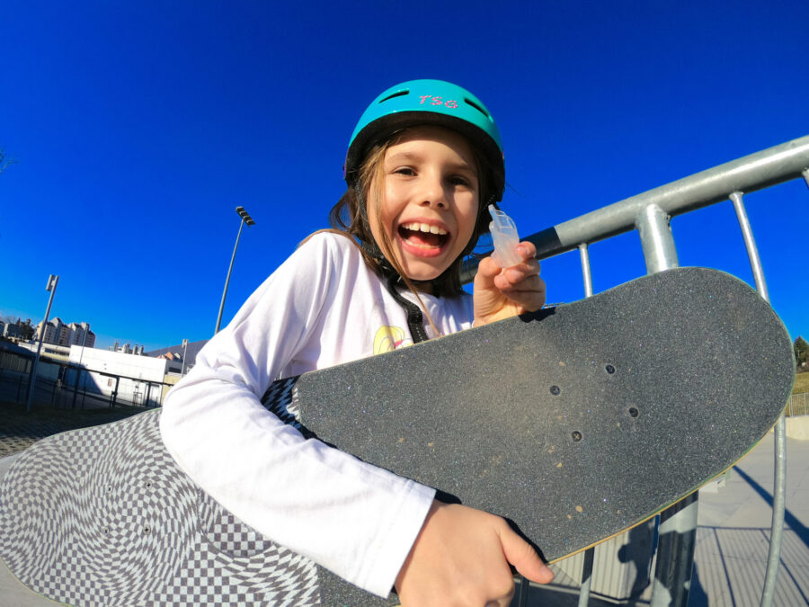 Young girl with helmet and skateboard smiles with her custom mouthguard to protect her teeth at the skatepark