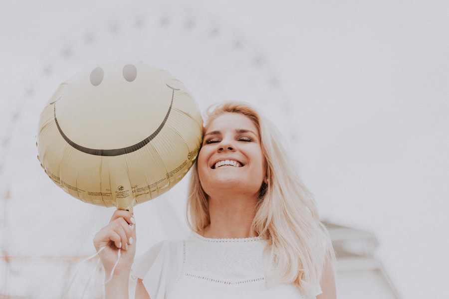 Blonde woman smiles as she holds a yellow smiley balloon with a Ferris wheel in the background