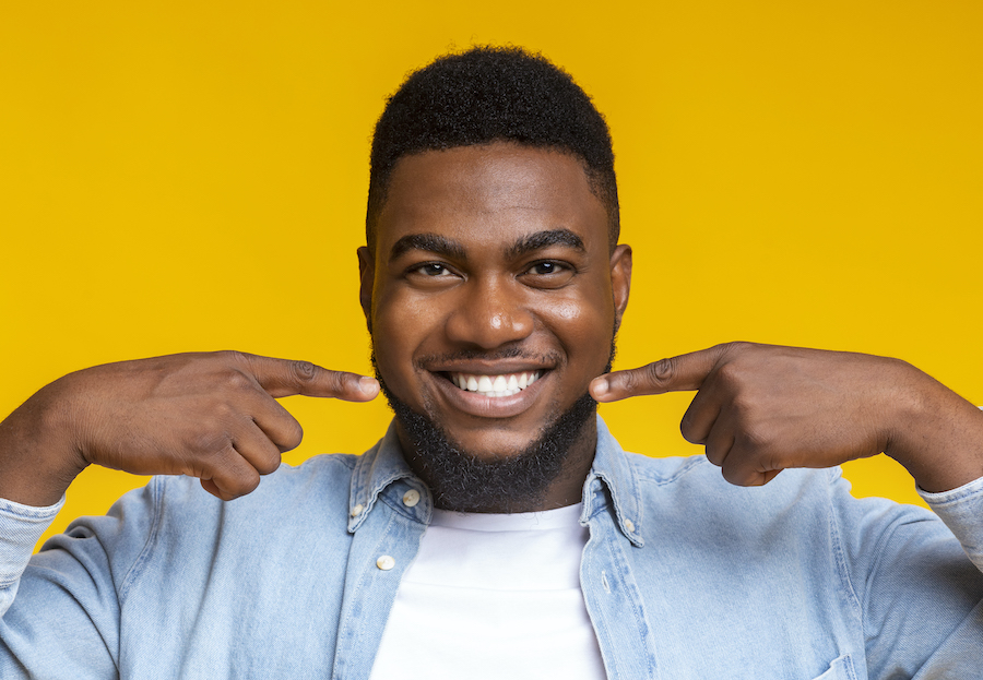 Closeup of a Black man in a white shirt smiling and pointing to his teeth against a yellow background