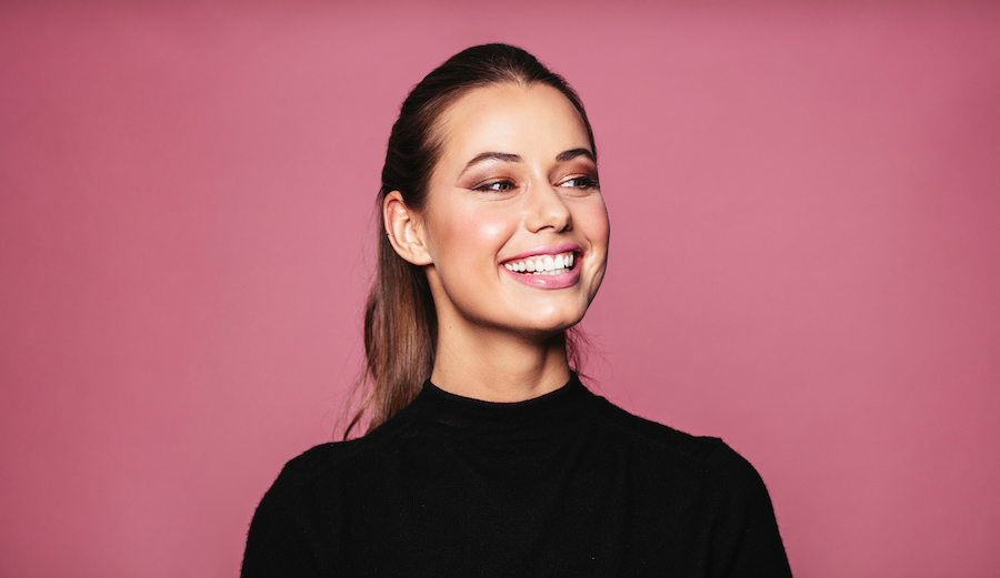 Brunette woman in a black blouse smiles brightly against a pink background after receiving BOTOX injections