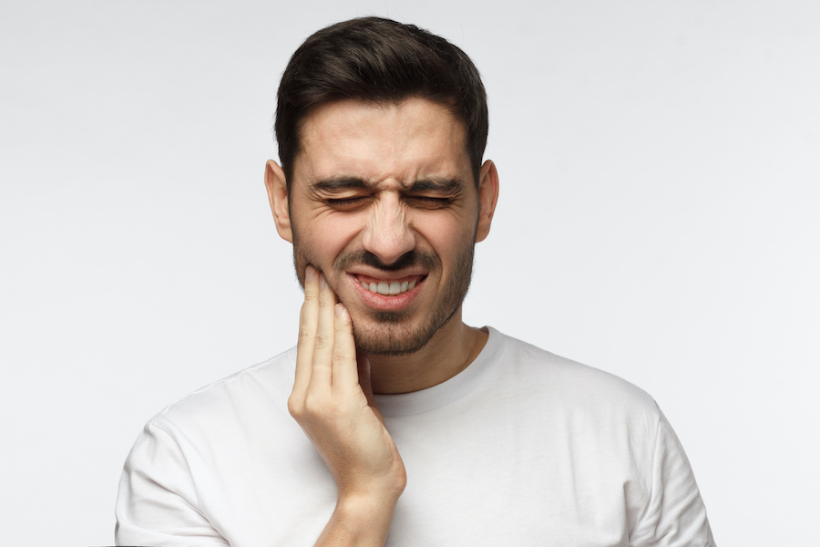 Closeup of a brunette young man in a white t-shirt cringing in pain and touching his cheek due to tooth pain